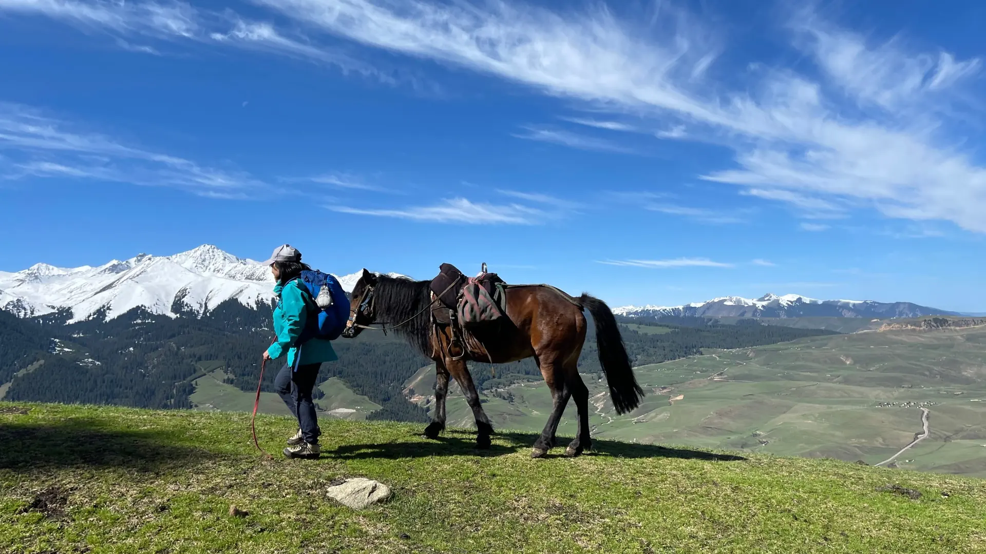 A trekker and a horse on a grassy plateau facing snow-capped mountains, Xinjiang, China